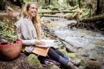 Young woman relaxing on forest riverbank with notebook — Stock Photo
