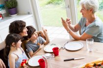 Grand-mère assise à la cuisine, photographiant sa fille adulte et ses petits-enfants, à l'aide d'un smartphone — Photo de stock