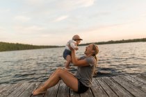 Mother sitting on lake pier holding up baby daughter — Stock Photo