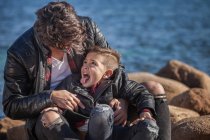 Father and son outdoors, fooling around, sitting on rocks beside sea — Stock Photo