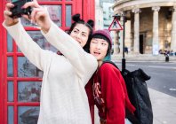 Two young stylish women taking selfie by red phone box, London, UK — Stock Photo