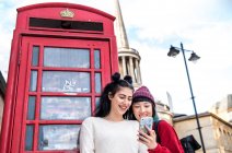 Two young stylish women looking at smartphone by red phone box, London, UK — Stock Photo