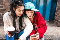 Дві молоді жінки сидять на тротуарі і дивляться на смартфон — стокове фото