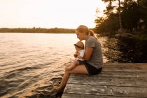 Mother sitting on pier with baby daughter looking down at  lake — Stock Photo