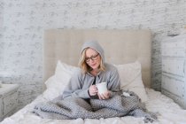 Молода жінка, загорнута в ковдру, сидить на ліжку з чашкою кави — стокове фото