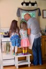 Вид сзади на девочку и сестру на стуле, смотрящих, как отец готовит еду на кухне — стоковое фото