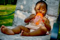 Baby girl sitting on garden lounge chair drinking from baby cup — Stock Photo