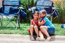 Due ragazze sedute sul marciapiede, una ragazza che sussurra all'orecchio degli amici — Foto stock