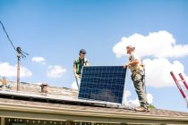 Два рабочих готовятся установить солнечную панель на крыше дома, вид с низкого угла — стоковое фото