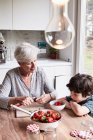 Grand-mère assise à la table de la cuisine, préparant des fraises, petit-fils assis à côté d'elle, regardant — Photo de stock