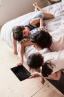 Madre, hijo e hija acostados en la cama, usando tableta digital, vista elevada - foto de stock