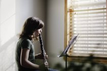 Chica con clarinete y soporte de música práctica por ventana - foto de stock