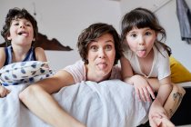 Retrato de madre, hijo e hija, acostados en la cama, sacando la lengua - foto de stock