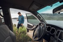 Mid adult man standing beside Dillon Reservoir, holding smartphone, view through parked car, Silverthorne, Colorado, USA — Stock Photo