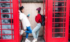Two young stylish women leaning against red phone boxes, London, UK — Stock Photo