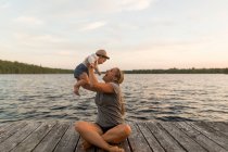 Mother sitting cross legged on lake pier holding up baby daughter — Stock Photo