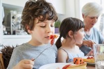 Young boy eating lunch at table with sister and grandmother — Stock Photo