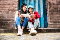 Дві молоді жінки сидять на тротуарі і дивляться на смартфон — стокове фото