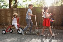 Three children in mini parade, banging drum, riding tricycle and using scooter — Stock Photo