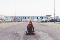 Red haired woman sitting with cross legged in car park — Stock Photo