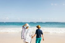 Madre e hijo, cogidos de la mano, de pie en la playa, mirando a la vista, vista trasera - foto de stock