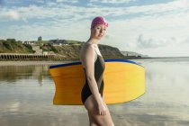 Молодая женщина, несущая доску для серфинга на берегу, смотрит в камеру — стоковое фото