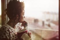 Jeune femme par fenêtre tenant boisson chaude — Photo de stock