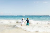 Madre e hijo, cogidos de la mano, de pie en el surf en la playa, vista trasera - foto de stock