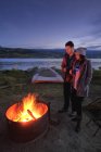 Couple debout près du feu de camp avec des boissons chaudes au crépuscule — Photo de stock