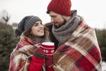 Romántica pareja joven envuelta en manta mientras compraba árbol de Navidad en el bosque - foto de stock
