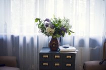Vase with bouquet of flowers on bedside table by window — Stock Photo