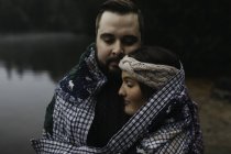Casal envolto em cobertor abraçando pelo lago — Fotografia de Stock