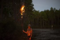 Молодий чоловік тримає гілку палаючого дерева, стоячи біля води в сутінках — стокове фото