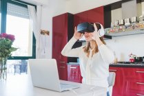 Молодая женщина за кухонным столом, просматривающая гарнитуру виртуальной реальности — стоковое фото