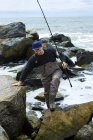 Joven pescador macho trepando por las rocas de la playa - foto de stock