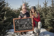 Fille et mère dans la forêt d'arbres de Noël avec signe de Noël joyeux, portrait — Photo de stock