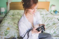 Молодая женщина сидит на кровати, рассматривая цифровую камеру — стоковое фото