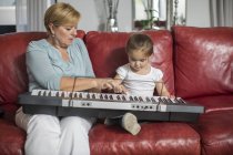 Girl and grandmother playing keyboard on red sofa at home — Stock Photo
