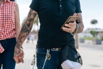 Pareja hipster tatuada cogida de la mano y el teléfono inteligente, sección media - foto de stock