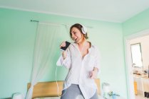 Jeune femme sur le lit portant des écouteurs et chantant dans le smartphone — Photo de stock