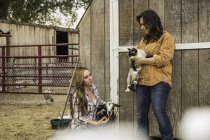 Mujer joven y madre sosteniendo una cabra y un gato en la cerca del rancho, Bridger, Montana, EE.UU. - foto de stock