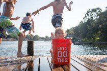 Young girl hiding behind bucket on jetty, children jumping into lake — Stock Photo