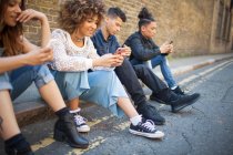 Quatre amis assis dans la rue, regardant les smartphones — Photo de stock