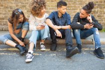 Quatre amis assis dans la rue, regardant les smartphones — Photo de stock