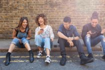 Четверо друзей сидят на улице, смеются, молодая женщина держит смартфон — стоковое фото