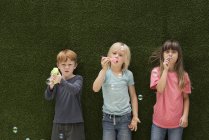 Crianças na frente da parede de grama artificial soprando bolhas — Fotografia de Stock