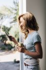 Счастливая молодая женщина у двери патио смотрит в смартфон — стоковое фото