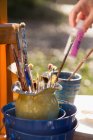 Pinceles en jarra y niña seleccionando pintura en el jardín, primer plano de la mano - foto de stock