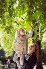 Mother holding up toddler daughter to touching leaves in park — Stock Photo