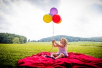 Girl sitting on red blanket in rural field looking up at bunch of colourful balloons — Stock Photo